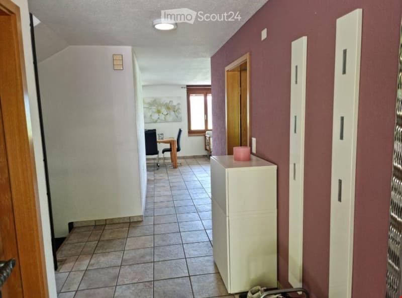A narrow hallway with tiled flooring, white walls, and a light fixture on the ceiling. A small table and chairs are visible in an adjacent room.