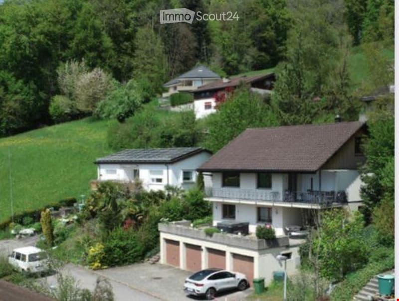 Aerial view of a two-story house with a garage, surrounded by greenery and other houses on a hillside.