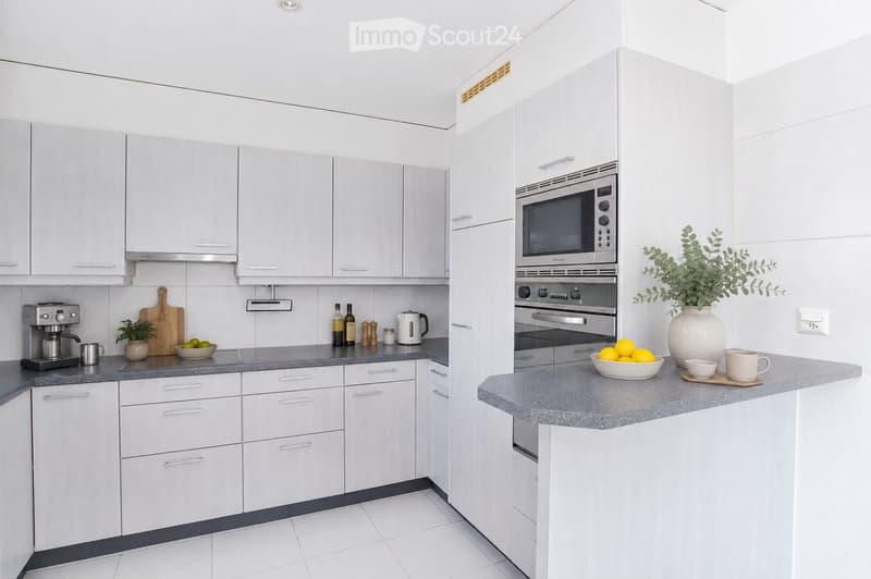 Modern kitchen with white cabinets, stainless steel appliances, and a grey countertop. Features a coffee maker, cutting board, bowl, oven, microwave, and decorative items including a vase with greenery and a bowl of lemons.