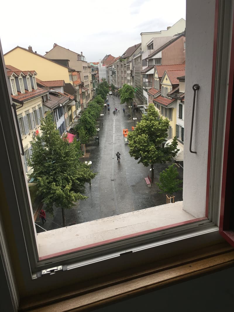 View from a balcony overlooking a wet, cobblestone street lined with colorful buildings, trees, and pedestrians. The street is wet from recent rain, and there are benches and bicycles visible.