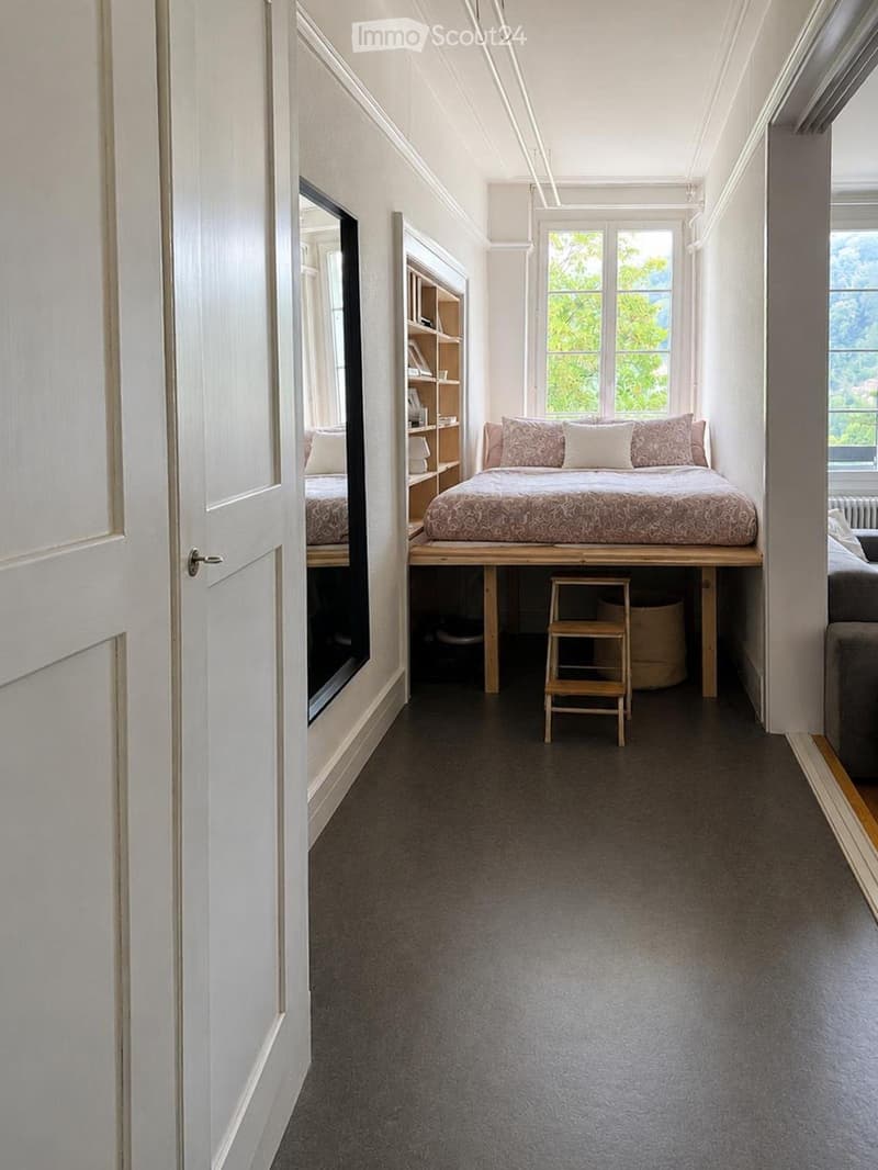 Cozy bedroom with a raised platform bed, pillows, a small wooden stool, a bookshelf, and a large mirror on the wall.