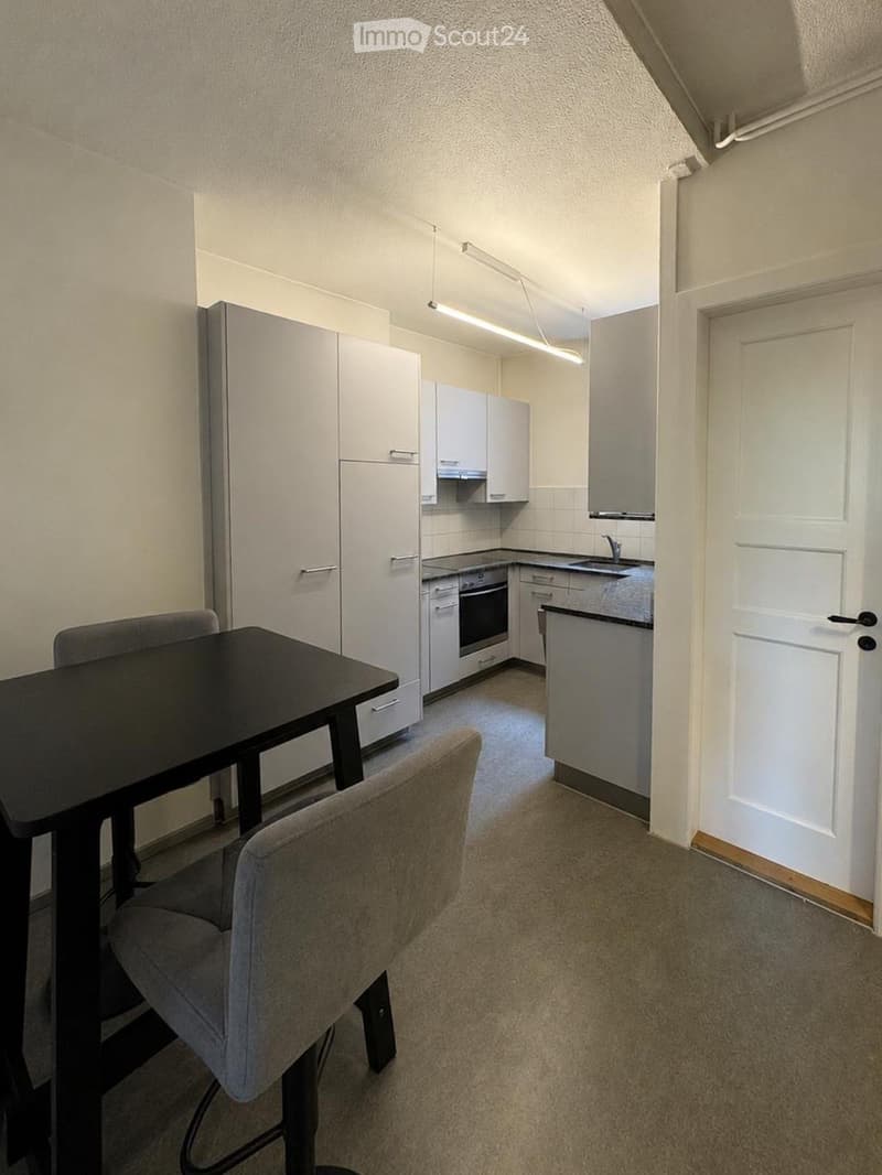A small, modern kitchen with white cabinets, a black dining table, and a grey chair. The kitchen features a sink, oven, and refrigerator.