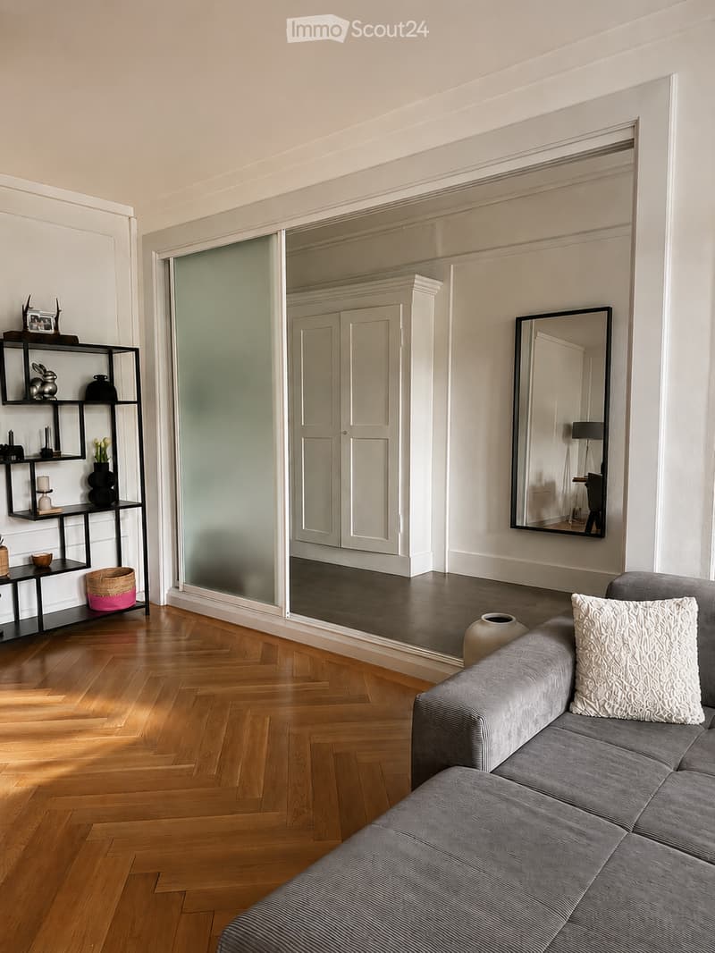 Modern living room with wooden herringbone flooring, grey sofa with decorative pillow, black shelving unit with various decorative items, large mirror, and a glass door leading to another room.