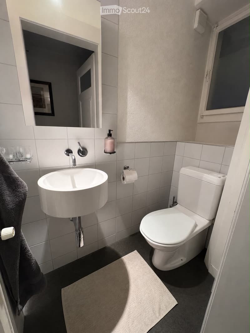 Modern bathroom with white tiles, a sink, toilet, soap dispenser, and a towel on the side.
