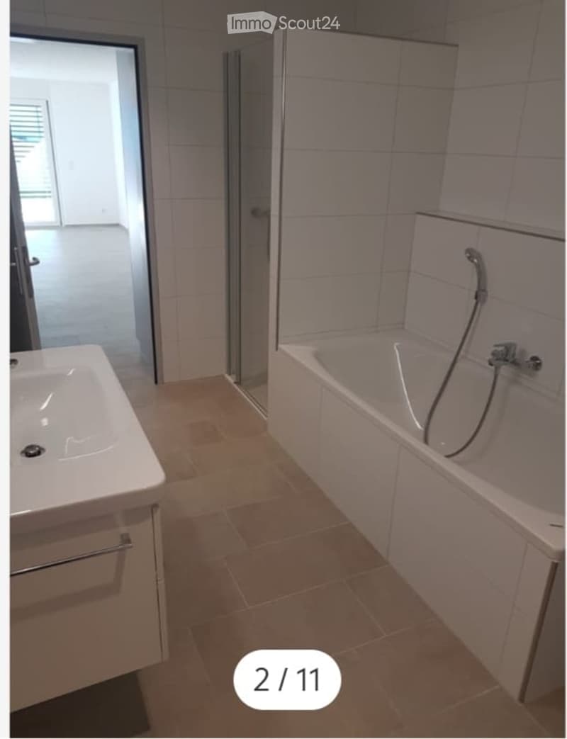 Modern bathroom with a white bathtub, a sink with a faucet, and a shower hose attached to the wall. The floor is tiled in a warm beige color, and the walls are covered with white tiles.