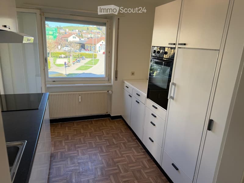 Modern kitchen with white cabinets, black countertop, and a built-in oven. The room features wooden herringbone flooring and a large window offering a view of the street.