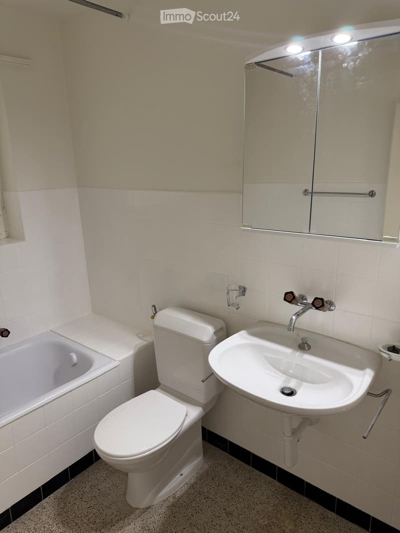 Modern bathroom with white tiles, a toilet, a sink, and a bathtub, featuring a large mirror with built-in lighting above the sink.