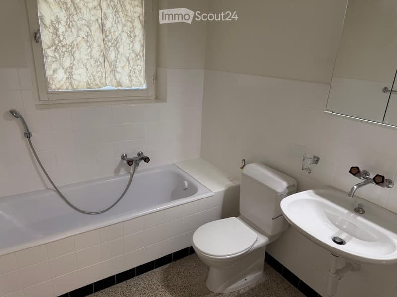 Bathroom with white tiled walls, a bathtub with a handheld shower, a toilet, and a sink with a mirror above it. The window has a frosted glass cover.
