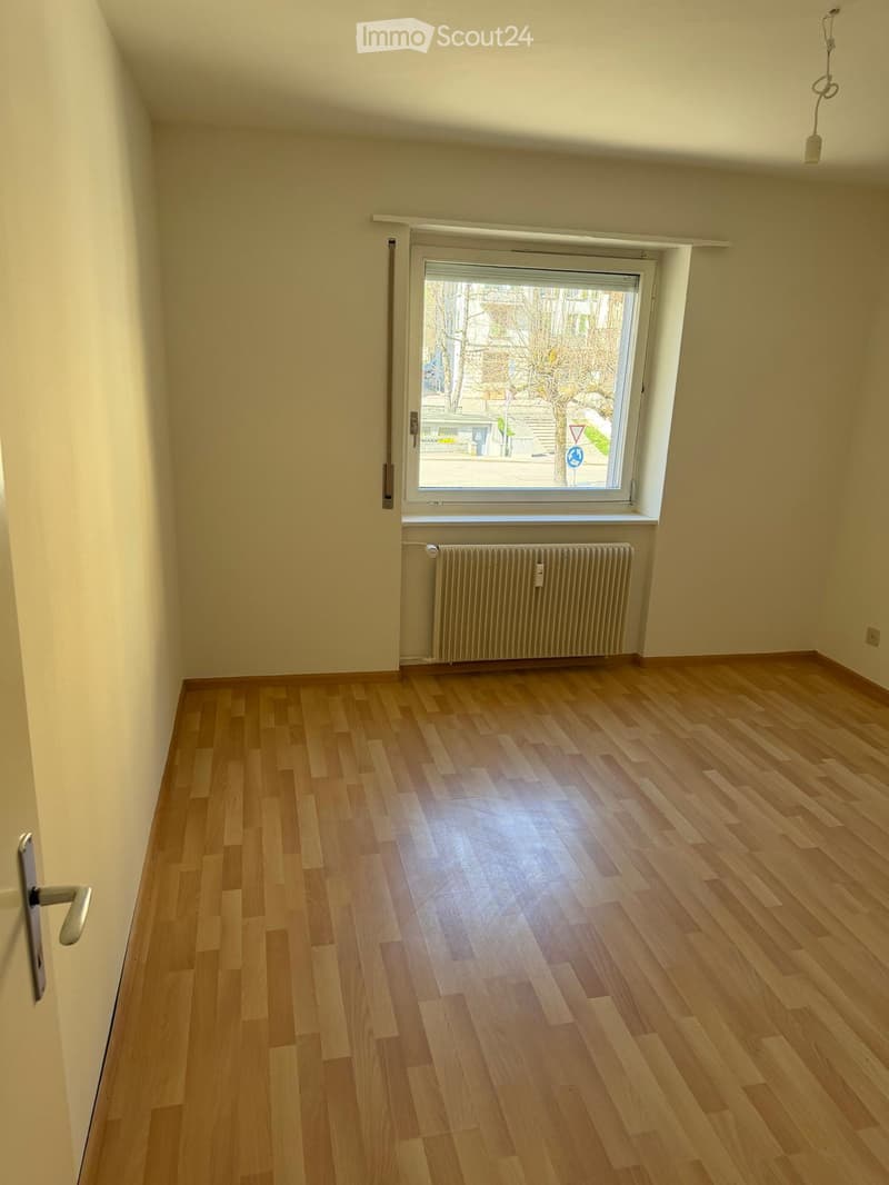 Empty room with light wooden flooring, white walls, a window with blinds, and a radiator below the window.