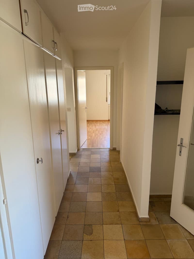 A clean, well-lit hallway with tiled flooring and white walls. Doors line both sides, leading to other rooms. The hallway has a simple, modern design with neutral tones.