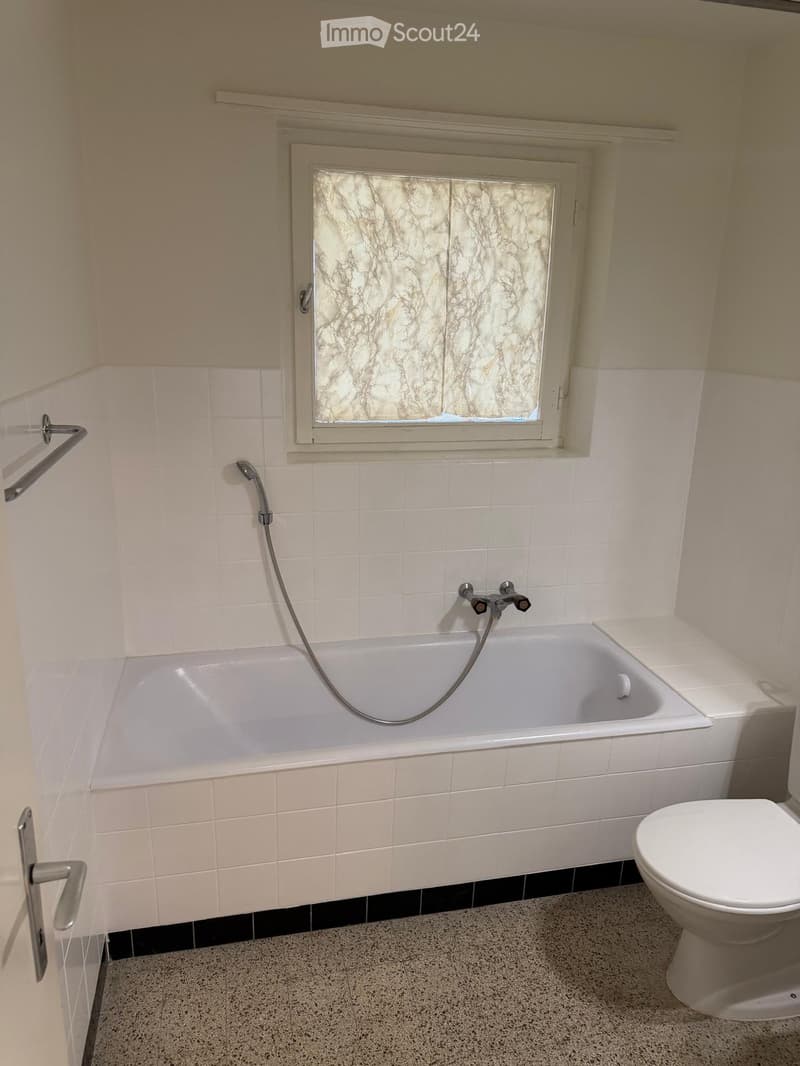 Bathroom with a bathtub, shower hose, toilet, window, and door handle.
