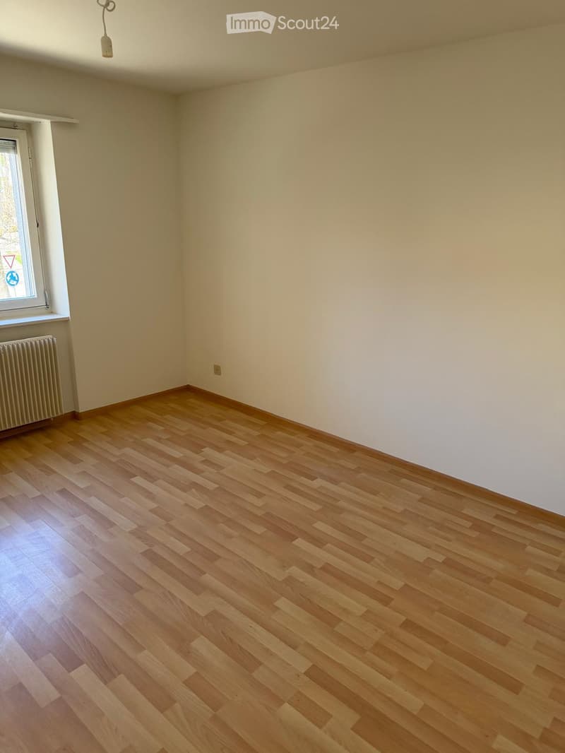 Empty room with light wooden flooring, white walls, and a single window with a radiator below it.