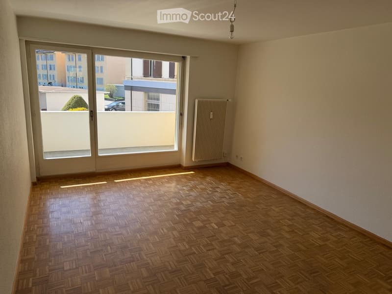 Empty room with wooden parquet flooring, large sliding glass doors leading to a balcony, radiator on one wall, and natural light streaming in.