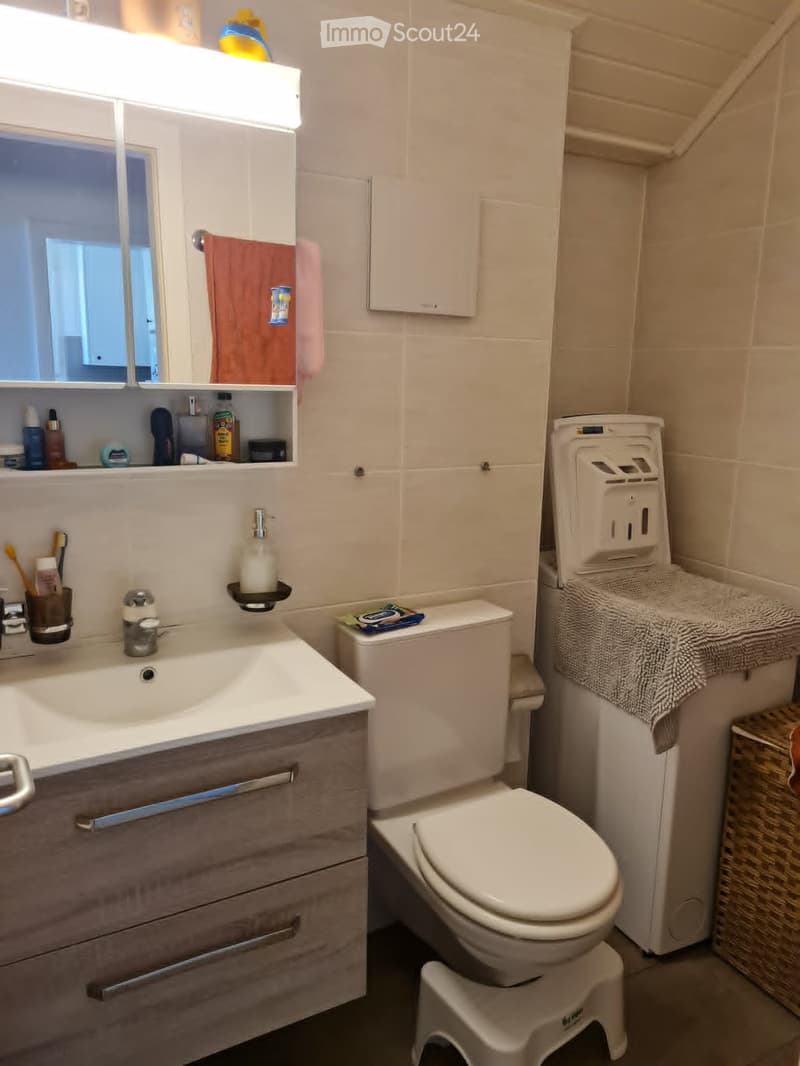 Modern bathroom with a white sink, mirror, and toilet. Features a wooden vanity with drawers, a towel hanging on the wall, and a small stool in front of the toilet.