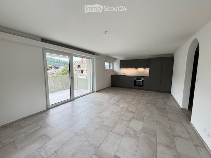 Bright, open-plan living and dining area with large sliding glass doors leading to a balcony, modern kitchen with dark cabinets, oven, and sink, tiled floor, and scenic mountain view.