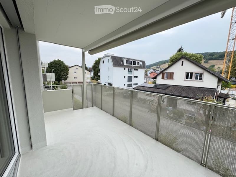 Balcony with a modern glass railing, overlooking a residential area with houses and a construction crane in the distance.