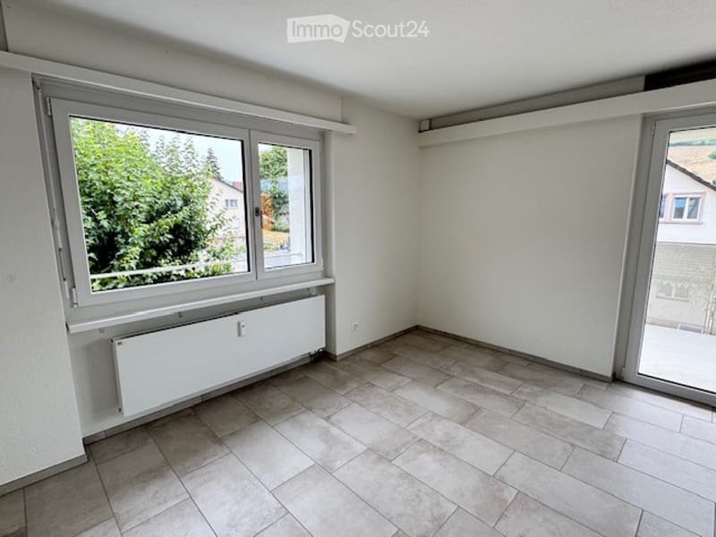 Empty room with white walls, tiled floor, large window with view of greenery, radiator, and sliding glass door leading outside.