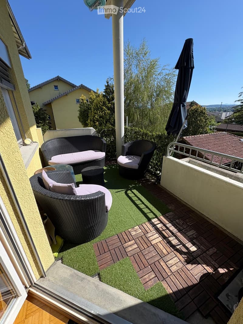 Balcony with wicker furniture, including a sofa, chairs, a small table, and a closed umbrella, set on artificial grass and wooden decking.
