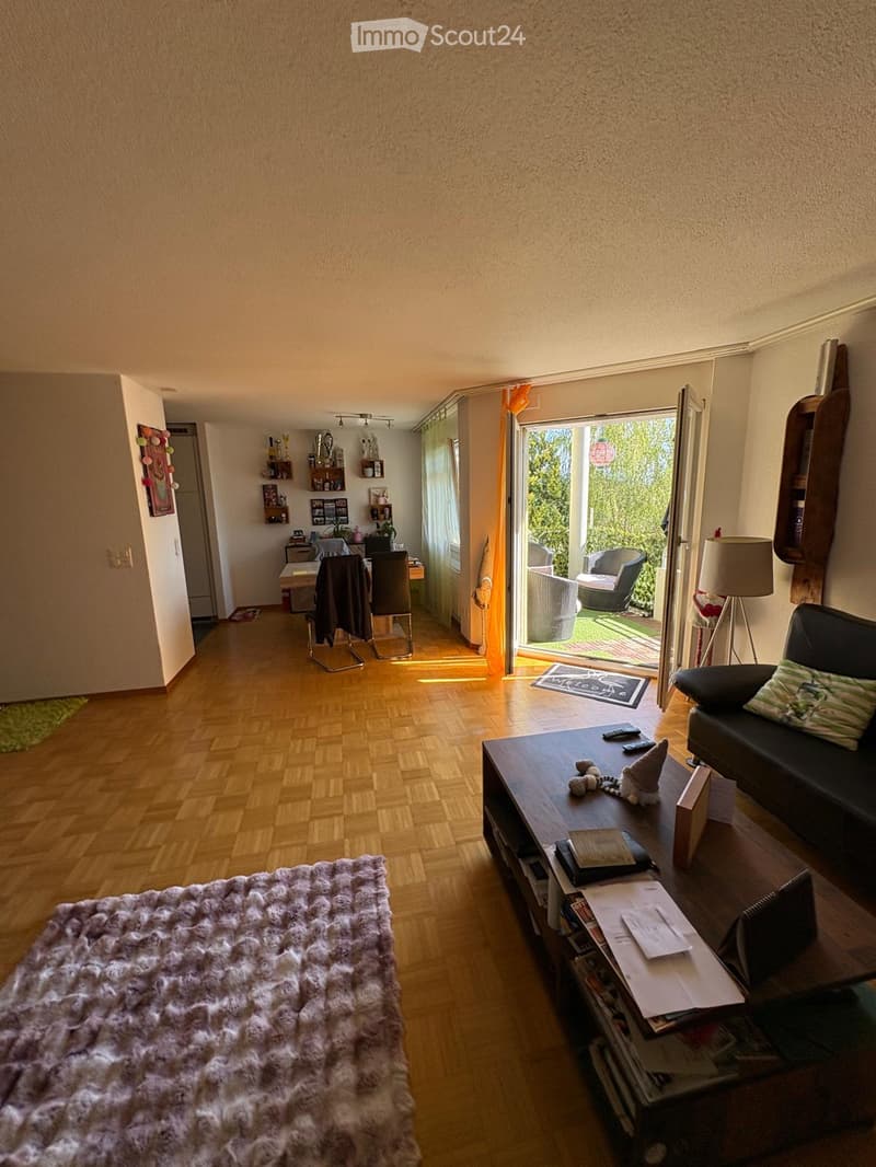 Living room with wooden flooring, a large sofa, coffee table, and a dining area with chairs and a table. There is a balcony with outdoor seating visible through the open sliding doors.