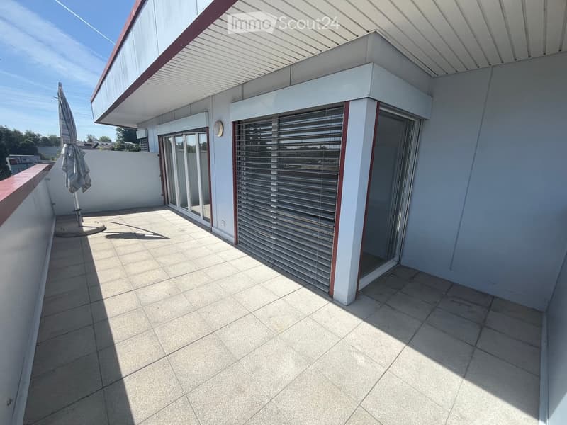 Modern balcony with tiled floor, sliding glass doors, and a closed shutter. Features a folded umbrella and a small wall-mounted light fixture.