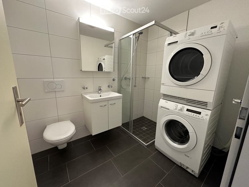 Modern bathroom with white tiles, a toilet, sink, mirror, glass-enclosed shower, and stacked washing machine and dryer.