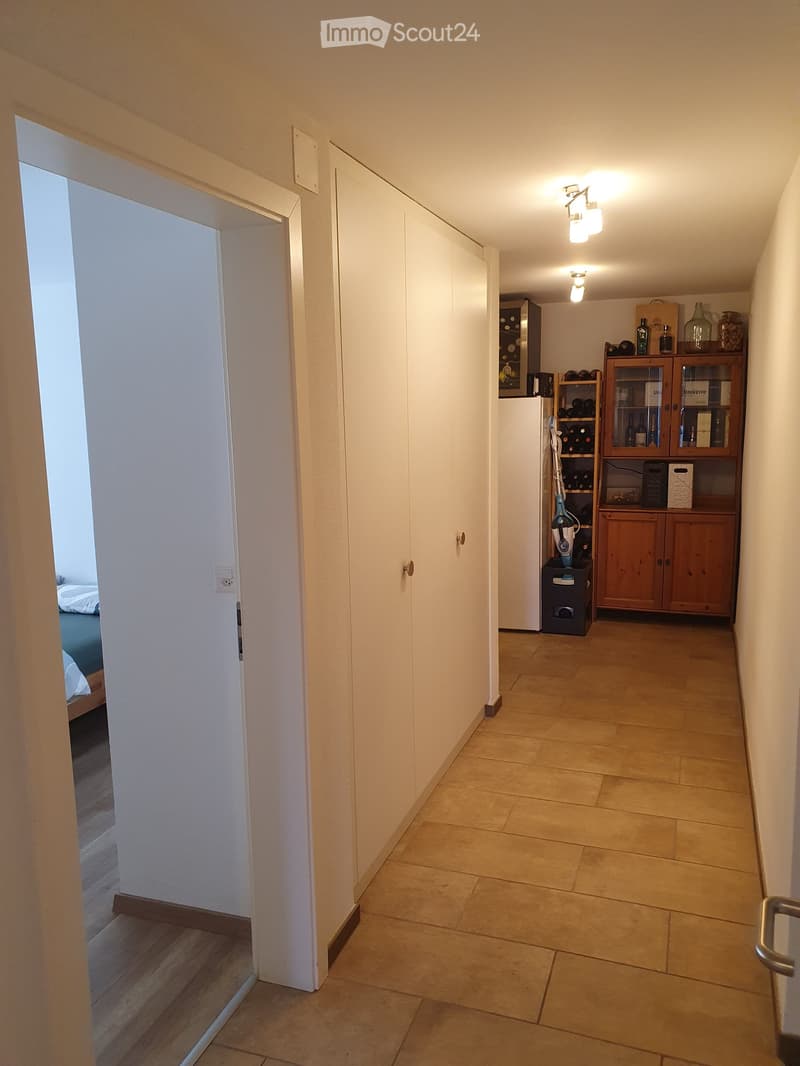 A well-lit hallway with tiled flooring, white walls, and built-in storage units. A door leads to a bedroom, and a small area with shelves and a refrigerator is visible at the end.
