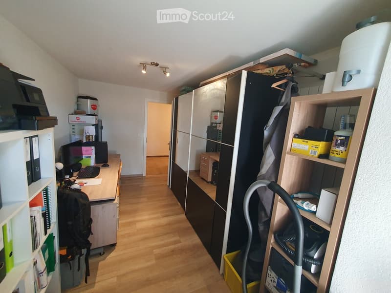 A furnished room with a wooden floor, a desk with a laptop and keyboard, a chair, a backpack, shelves, a large wardrobe, and a vacuum cleaner on a shelf.