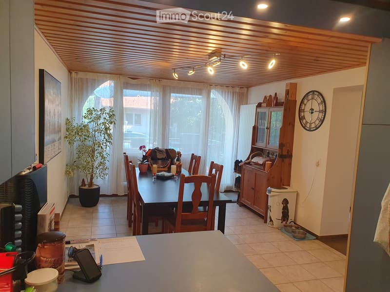 Dining area with a wooden table, chairs, a cabinet, a potted plant, and a clock on the wall. The room has a tiled floor and a wooden ceiling with recessed lighting.
