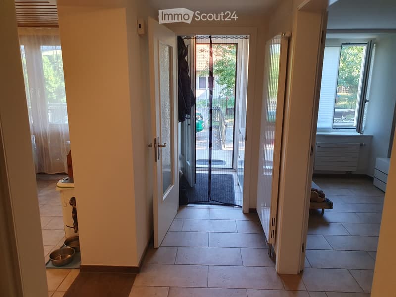 Hallway with tiled flooring, leading to an open door that reveals a balcony area with outdoor furniture and a view of greenery. There are pet bowls and a trash bin visible on the left side.