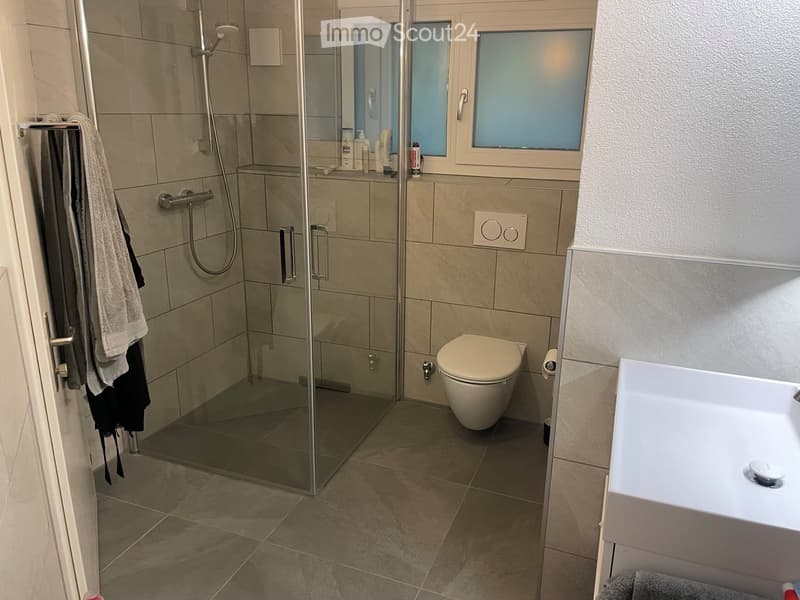Modern bathroom with a glass-enclosed shower, toilet, bathtub, and towel rack. The shower has a handheld sprayer and a built-in seat. The walls are tiled in beige, and the floor is tiled in a light brown color.