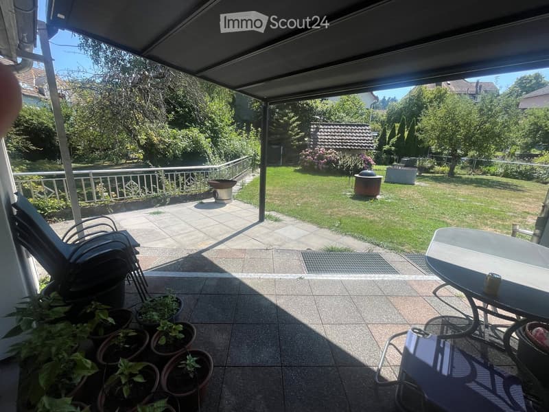 Balcony with tiled floor, potted plants, a table, and a chair, overlooking a well-maintained garden with a barbecue and flowers.