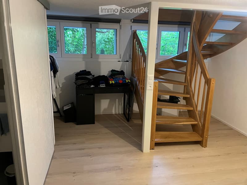 Bright hallway with wooden flooring, a staircase leading upwards, and a small table with folded clothes and a bag. Windows on the left side provide natural light.