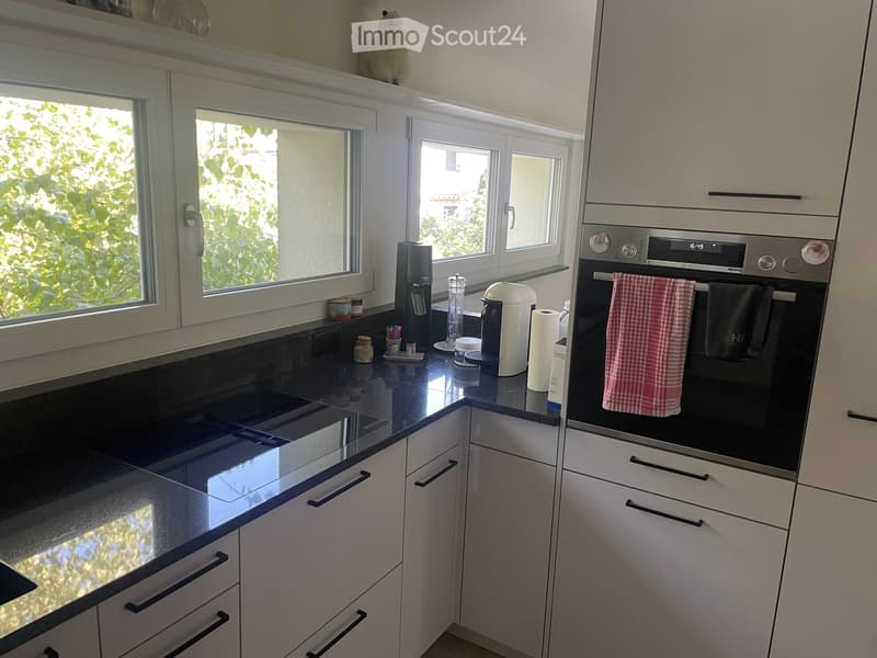 Modern kitchen with white cabinets, black countertops, and stainless steel appliances. Features a built-in oven with a digital display, a coffee maker, and various small kitchen items on the counter.