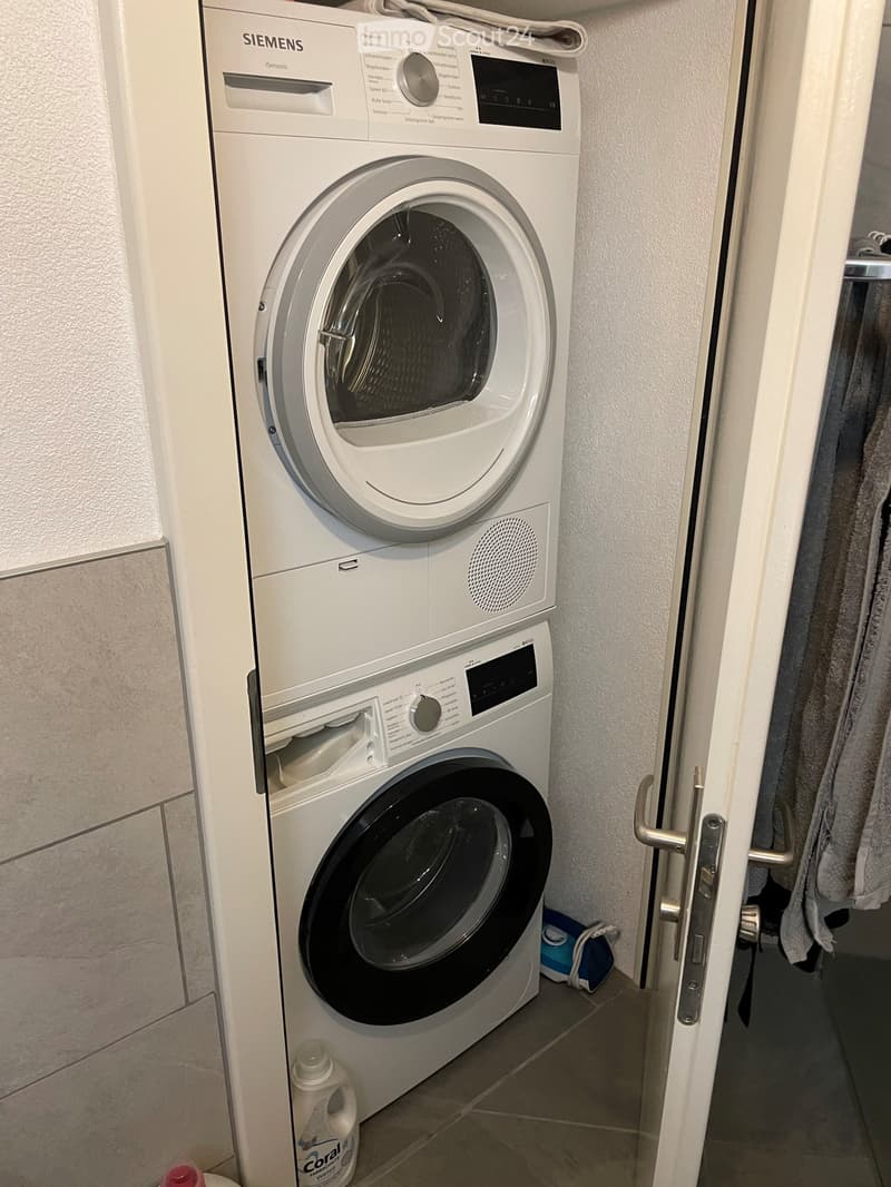 A compact bathroom with a stacked washing machine and dishwasher unit, white tiled walls, and a small shelf with cleaning supplies.