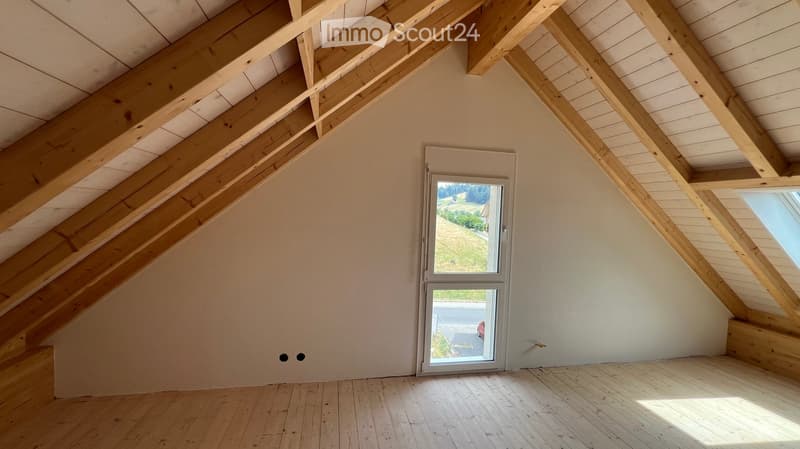 Empty attic space with wooden beams, wooden floor, and a large window showing a view of the outdoors.