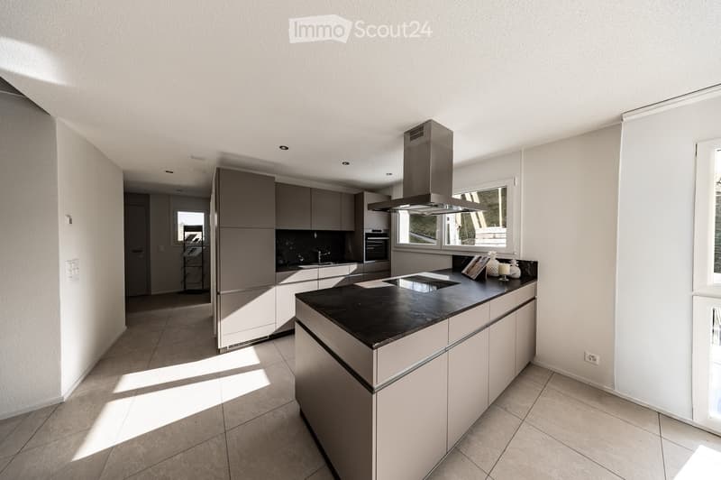 Modern kitchen with light grey cabinets, black countertop, island, built-in oven, and range hood. Natural light from large windows.