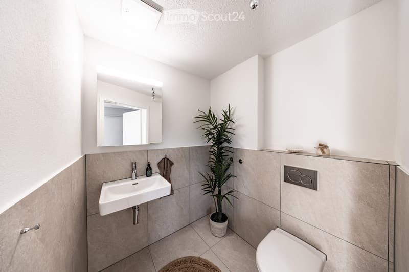 Modern bathroom with a white sink, toilet, mirror, and a potted plant. The walls are tiled in a neutral color, and the floor is tiled as well. A towel hangs on the wall, and a small decorative item sits on the counter.