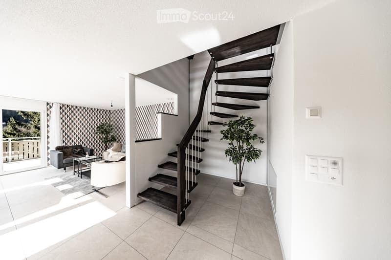 Modern living room with a spiral staircase leading to an upper level, featuring a grey sofa, white armchair, coffee table, and potted plant.