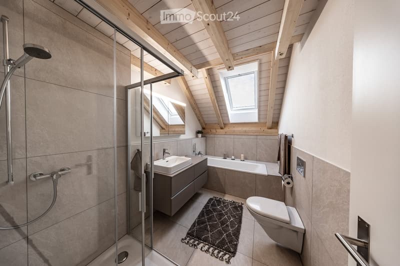 Modern bathroom with a glass-enclosed shower, a sink with a dark wooden cabinet, a bathtub, a toilet, and a small rug on the floor. The ceiling has exposed wooden beams and skylights.