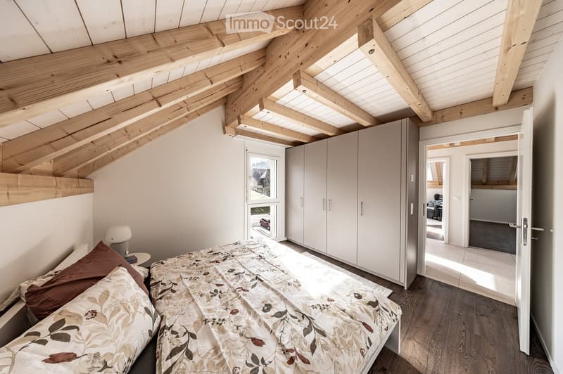 Bright bedroom with wooden slanted ceiling beams, large window, bed with patterned bedding, nightstand with lamp, and built-in wardrobe.