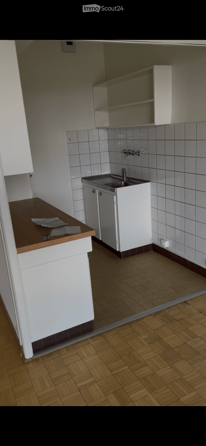 Modern kitchen with white cabinets, a stainless steel sink, and a wooden countertop. The floor is made of light brown wood tiles, and the walls are tiled with gray tiles.