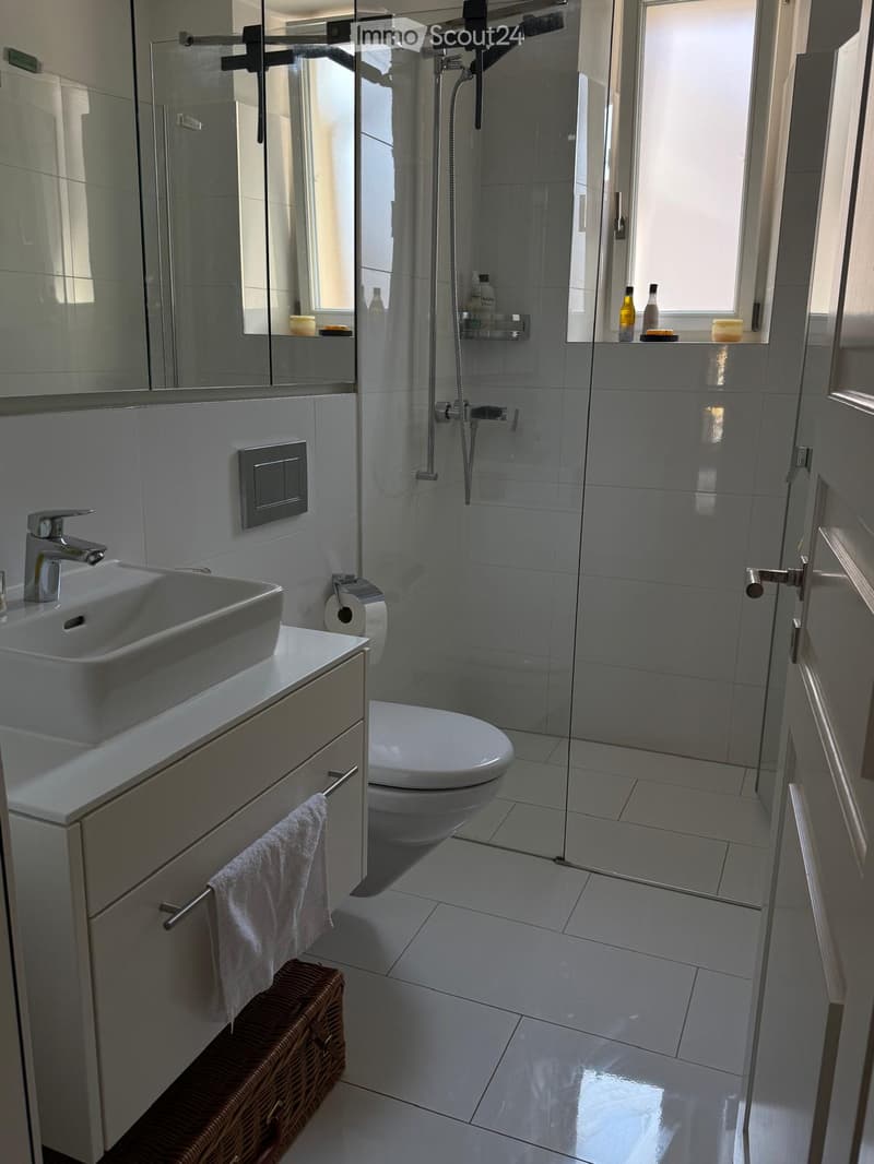 Modern bathroom with white tiles, a sink with a modern faucet, a toilet, a glass-enclosed shower, and a large mirror. A white towel hangs on a rack, and a wicker basket is placed on the floor.