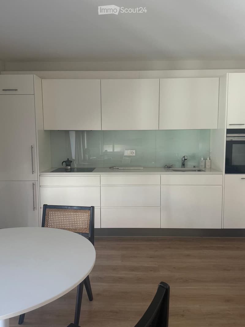 Modern kitchen with white cabinets, a round white dining table, and a wicker chair. The kitchen features a sink, faucet, oven, and coffee maker on the counter.