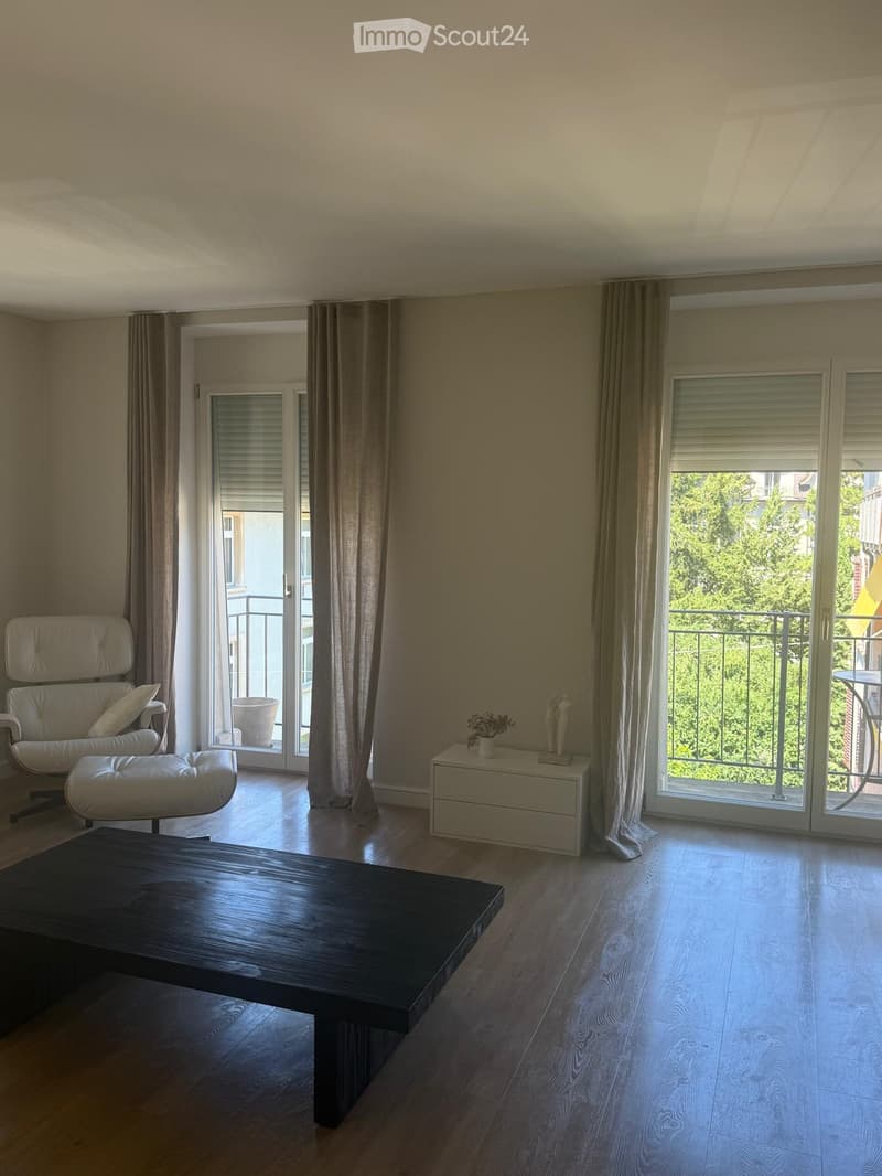 Modern living room with wooden flooring, large windows, and a balcony. Features a sofa, coffee table, side table, and a small table on the balcony.