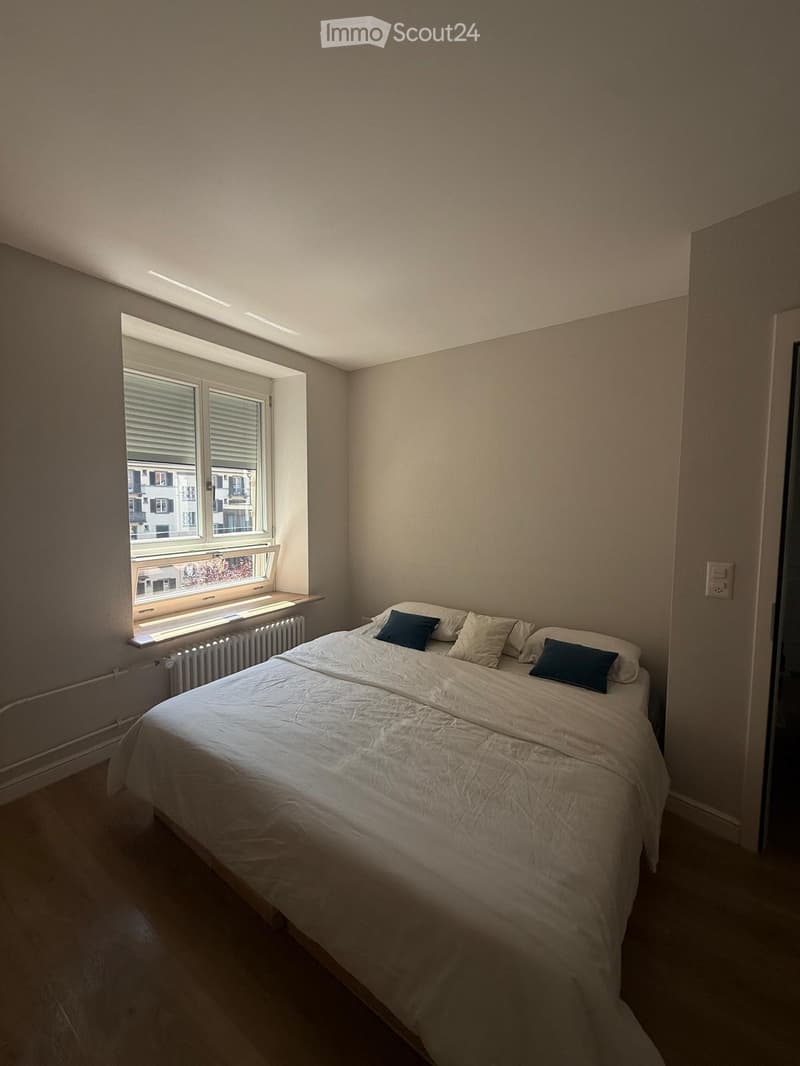 A small, neatly arranged bedroom with a single bed, white bedding, and two blue pillows. The room features a large window with blinds, a radiator below it, and a simple, modern design with light-colored walls and flooring.