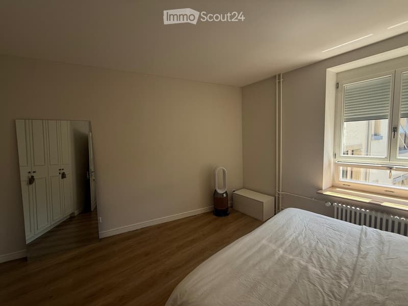 Bedroom with wooden flooring, a bed with white bedding, a wardrobe, a fan, and a radiator near the window.