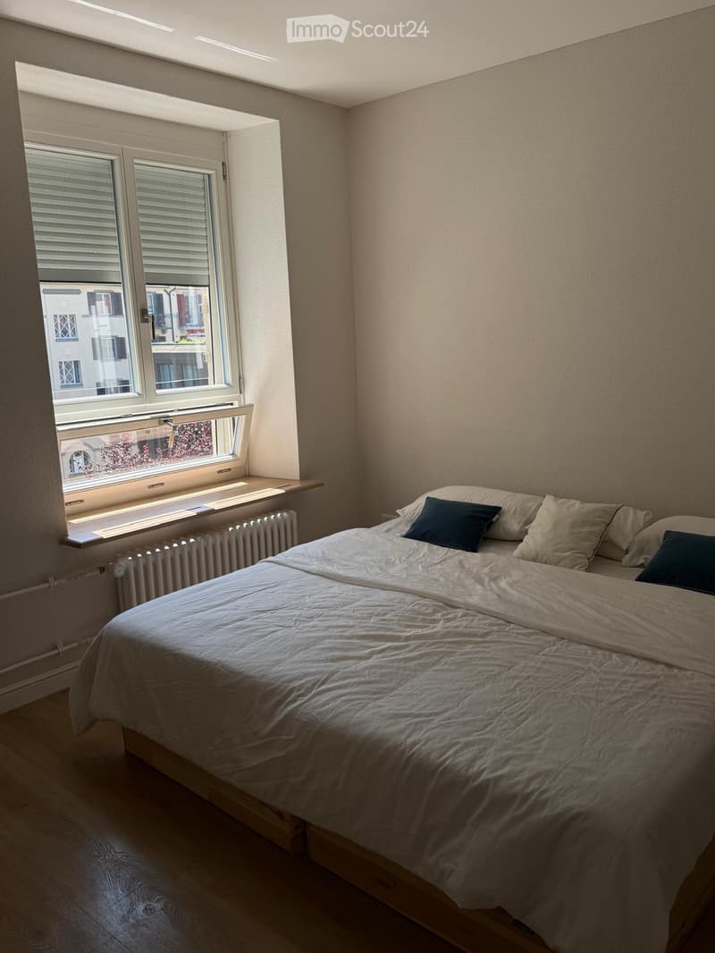 A tidy bedroom with a single bed, pillows, and a radiator beneath a large window with blinds.