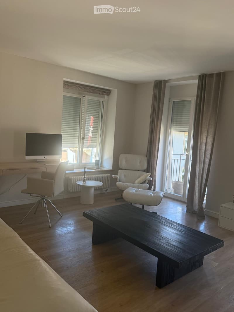 Modern living room with wooden flooring, large windows, balcony access, and contemporary furniture including a sofa, chairs, a coffee table, and a screen.