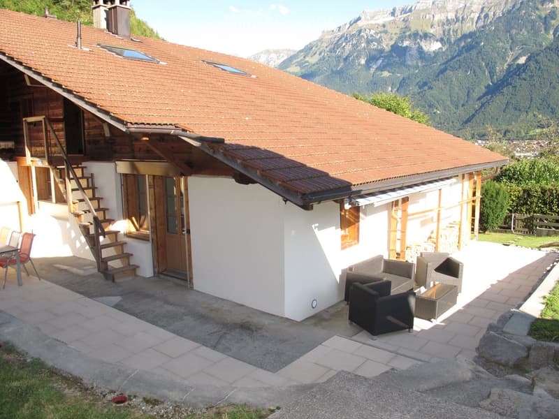 Two-story wooden house with a red-tiled roof, white walls, and a spacious patio area with outdoor furniture, overlooking a scenic mountain backdrop.