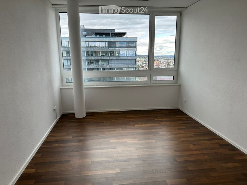 Empty room with wooden flooring, white walls, and large windows offering a view of a modern building and cityscape.